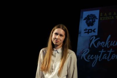 Dziewczyna recytuje dla publiczności. Wokół niej ciemność.
