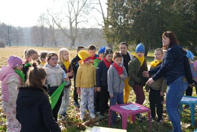 Grupa dzieci w parku stoi nieopodal stolików, przed nimi pani.