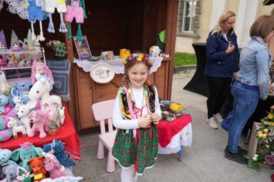 Dziewczynka ubrana na ludowo stojąca uśmiechnięta przed stoiskiem z dekoracjami.