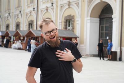 Uśmiechnięty pan pozuje do zdjęcia. Za nim stoiska na kiermaszu.