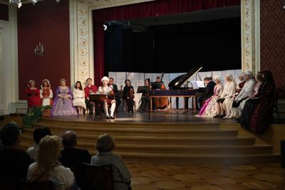 Osoby w historycznych przebraniach siedzą na scenie wokół artystów grających na instrumentach.