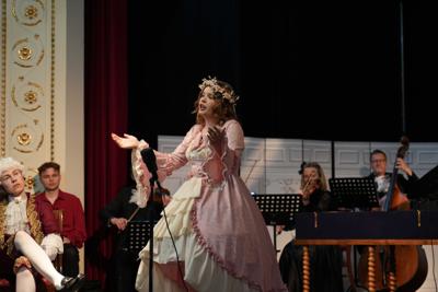 Osoby w historycznych przebraniach występują na scenie. Za nimi artyści grający na instrumentach.