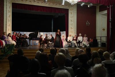 Osoby w historycznych przebraniach siedzący wokół artystów grających na instrumentach na scenie.