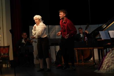 Artyści w historycznych przebraniach występują na scenie. Za nimi orkiestra.