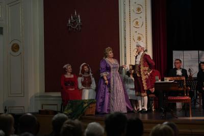 Artyści w historycznych przebraniach występują na scenie. Za nimi orkiestra.