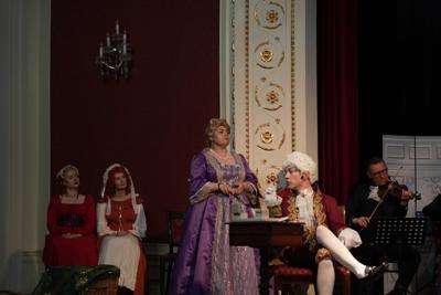 Artyści w historycznych przebraniach występują na scenie. Za nimi orkiestra.