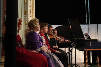 Artyści w historycznych przebraniach występują na scenie. Za nimi orkiestra.