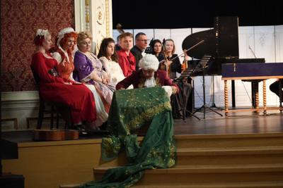 Artyści w historycznych przebraniach występują na scenie. Za nimi orkiestra.