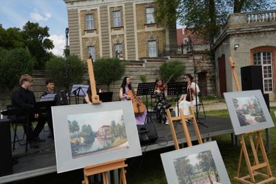 Grupa muzyków grająca koncert, przed sceną znajdują się sztalugi z obrazami.
