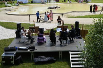 Grupa muzyków grająca koncert, przed sceną znajdują się sztalugi z obrazami. Przed nimi fontanna.