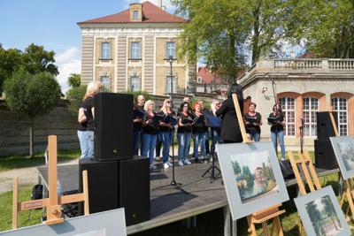 Występ chóru, przed sceną znajdują się sztalugi z obrazami. W tle budynek pałacu.