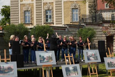 Występ chóru, przed sceną znajdują się sztalugi z obrazami. W tle budynek pałacu.