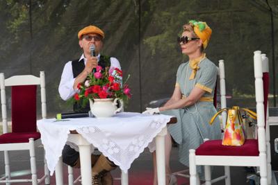 Staromodnie ubrani państwo siedzący przy stole. Pan mówi do mikrofonu.