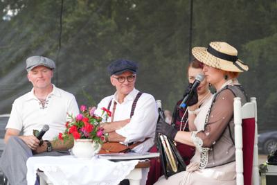 Grupa staromodnie ubranych osób siedzi przy stole z mikrofonami.