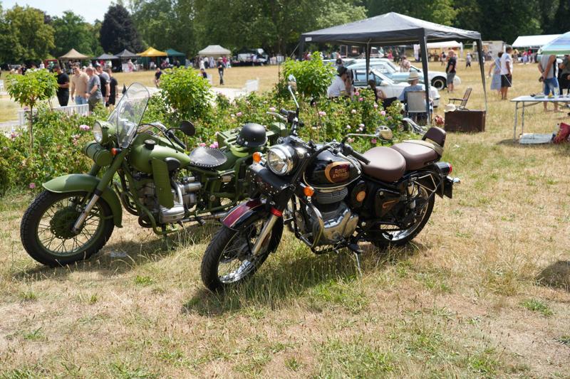 Staromodne motory. Za nimi krzewy. W oddali widać stoiska.