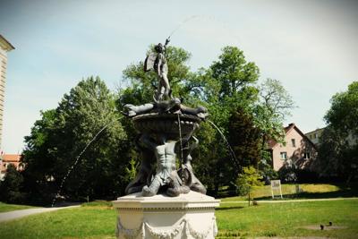 Park, na pierwszym planie fontanna. Na jej czubku postać ze skrzydłami i z widełkami.