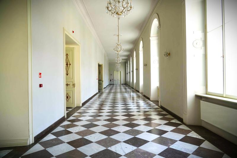 Długi korytarz. Na podłodze znajdują się czarno-białe kafelki. Na lewej ścianie znajdują się zdobione drzwi, na prawej okna. Na suficie żyrandole.