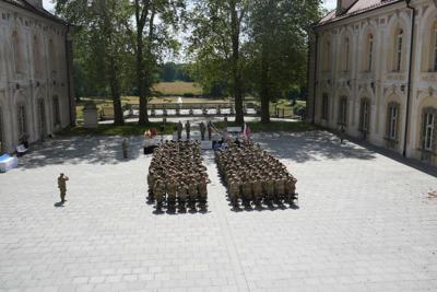 Grupa żołnierzy wstała z krzeseł by zasalutować. Znajdują się na dziedzińcu pałacu. W oddali widać drzewa, fontannę, park.