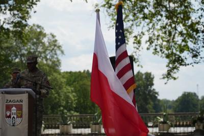 Żołnierz stoi przy mównicy. Obok flagi Polski i Stanów Zjednoczonych. W tle widać roślinność.