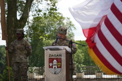 Żołnierz stoi przy mównicy, obok widać flagi, z tyłu drzewa.