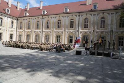 Grupa żołnierzy znajdująca się na placu otoczonym zabudową. Część siedzi na krzesłach, niektórzy znajdują się na scenie. Są także flagi.