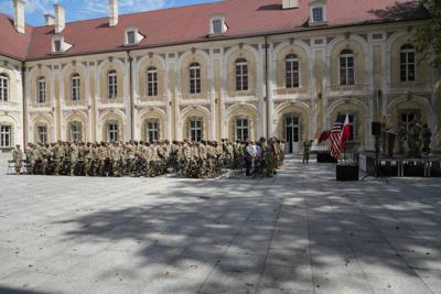 Grupa żołnierzy znajdująca się na placu otoczonym zabudową. Część siedzi na krzesłach, niektórzy znajdują się na scenie. Są także flagi.