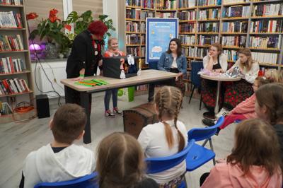 Zabawa w "Zgadnij co to jest?" z użyciem różnych przedmiotów przygotowanych przez Koło Gospodyń. W tle plakat z programem ferii w bibliotece.