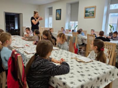 Pani prowadząca opowiada o karmnikach dla ptaków. Dzieci siedzą przy stolikach i słuchają jej. 