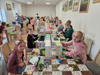 Dzieci siedzą przy stolikach, uśmiechają się i robią prace w pudełkach po butach  