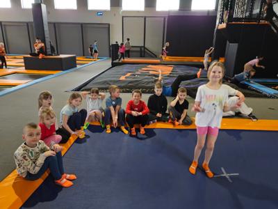 Dzieci siedzą na podłodze i uśmiechają się do zdjęcia. Dziewczynka na pierwszy planie stoi na trampolinie