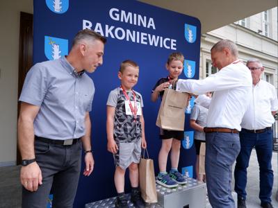 Krzysztof Krawczyk - przewodniczący Rady Miejskiej wręcza nagrodę stojącemu na podeście chłopcu za zajęcie 1 miejsca w biegach. Obok stoi Burmistrz Rakoniewic