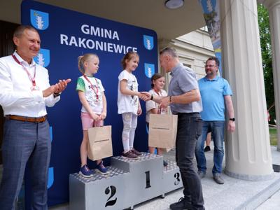Dr Gerard Tomiak - burmistrz gm. Rakoniewice gratuluje stojącej na podeście dziewczynce za zajęcie 1 miejsca w biegach. Obok stoi Arkadiusz Pawłowski oraz Krzysztof Krawczyk