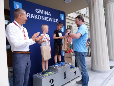 Arkadiusz Pawłowski - zastępca burmistrza gm. Rakoniewice wręcza nagrodę stojącemu na podeście chłopcu za zajęcie 1 miejsca w biegach. Obok stoi Krzysztof Krawczyk - przewodniczący rady Miejskiej