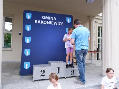 Arkadiusz Pawłowski - zastępca burmistrza gm. Rakoniewice wręcza medal stojącej na podeście dziewczynce za zajęcie 3 miejsca w biegach. 
