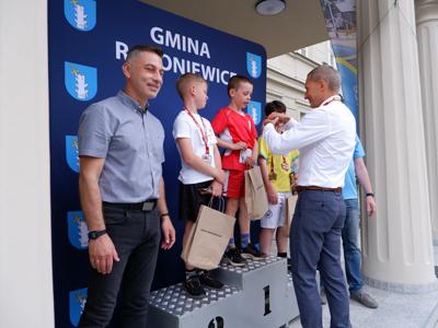 Krzysztof Krawczyk - przewodniczący Rady Miejskiej wręcza medal stojącemu na podeście chłopcu za zajęcie 1 miejsca w biegach. Obok stoi Burmistrz Rakoniewic