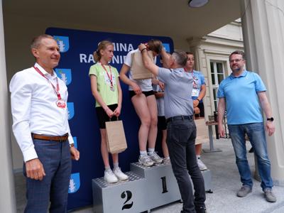 Dr Gerard Tomiak - burmistrz gm. Rakoniewice wręcza medal stojącej na podeście dziewczynie za zajęcie 1 miejsca w biegach. Obok stoi Arkadiusz Pawłowski i Krzysztof Krawczyk