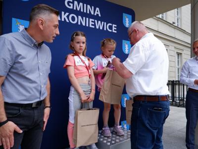 Marek Maćkowiak - zastępca przewodniczącego rady miejskiej wręcza nagrodę stojącej na podeście dziewczynce za zajęcie 1 miejsca w biegach. Obok stoi Burmistrz Rakoniewic 