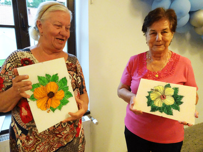 Dwie uśmiechnięte kobiety trzymające obrazki z kwiatami namalowane na szkle.