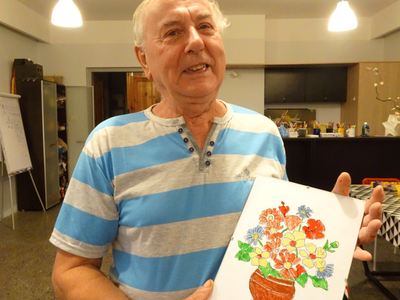 Uśmiechnięty mężczyzna trzymający kolorowy obrazek z kwiatami namalowanymi na szkle.