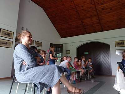 kobieta siedząca na krześle, za nią grupka dzieci w różnych pozach na krześle patrząca przed siebie