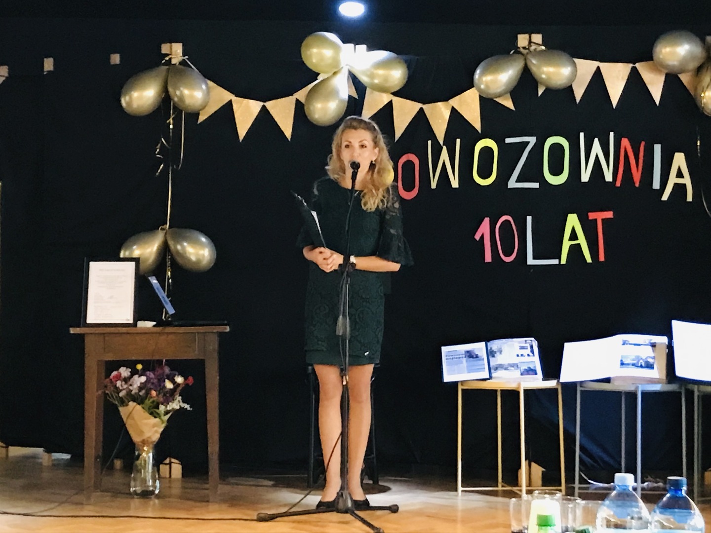 Zdjęcie kobiety w zielonej sukience, która mówi przemówienie przez mikrofon. W tle widać napis Powozownia 10 lat. 