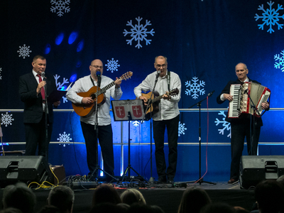 Fotografia wykonana w Zespole Szkół Ekonomiczno – Usługowych w Żychlinie. Przedstawia scenę na której stoją przedstawiciele gminy Sompolno: burmistrz Roman Bednarek (wokal), radny Andrzej Prętnicki (gitara), dyrektor MGOPS – Tomasz Majewski (gitara) oraz przedstawiciel seniorów z DDP – Zygmunt Teodorczyk (akordeon). Za nimi rozciąga się niebieska kotara z białymi śnieżynkami.