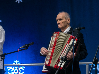Zdjęcie przedstawia starszego mężczyznę grającego na akordeonie. Przed nim mikrofony, za nim niebieskie tło.
