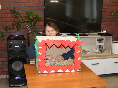 Zdjęcie wykonane w sali głównej Domu Dziennego Pobytu Senior+ w Sompolnie. Przedstawia kobietę, rozpoznającą za pomocą zmysłu dotyku to, co znajduje się w kartonie.