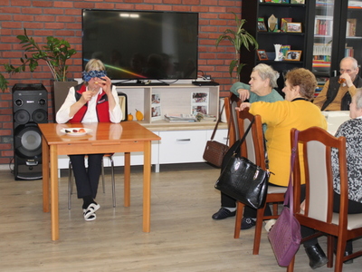 Zdjęcie przedstawia kobietę siedzącą przy stole z zawiązanymi oczami, rozpoznającą produkty spożywcze za pomocą zmysłu smaku. Po prawej stronie widać grupę seniorów siedzących przy stole.