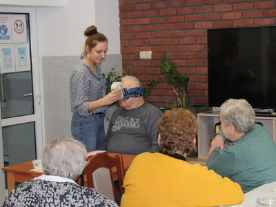Na zdjęciu widać mężczyznę siedzącego przy stole z zawiązanymi oczami. Obok niego stoi kobieta, która podstawia mu przyprawy do rozpoznania za pomocą zmysłu węchu.