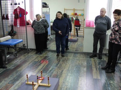 Na zdjęciu widać cztery osoby starsze grające w ringo na sali rehabilitacyjnej. W lustrze widać starszą kobietę ćwiczącą przy drabinkach.