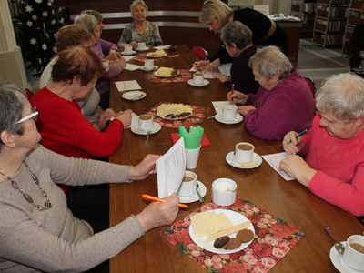 Na zdjęciu widać grupę seniorek siedzących przy stole i rozwiązujących test na kartkach.