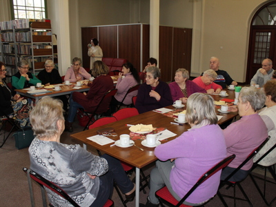 Na fotografii widać seniorów siedzących przy dwóch stołach. Wzrok seniorów skierowany jest na kobietę, która prowadzi wykład o zdrowym żywieniu. W tle regały z książkami.