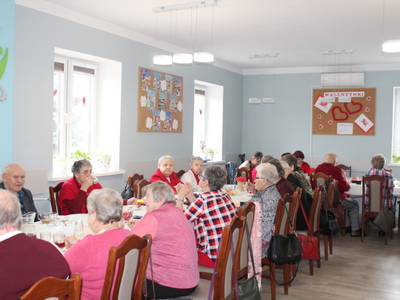 Fotografia wykonana w sali głównej Domu Dziennego Pobytu Senior+ w Sompolnie. Przedstawia uczestników siedzących przy stołach podczas balu karnawałowo – walentynkowego. Każdy z uczestników ma na sobie element ubioru w kolorze czerwonym.